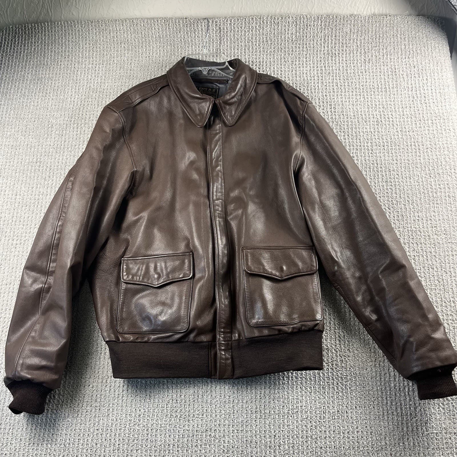 灰色の布の上に置いた、こげ茶の革のジャケットの前面。肩章と雨ぶたの付いた2つのポケットがある