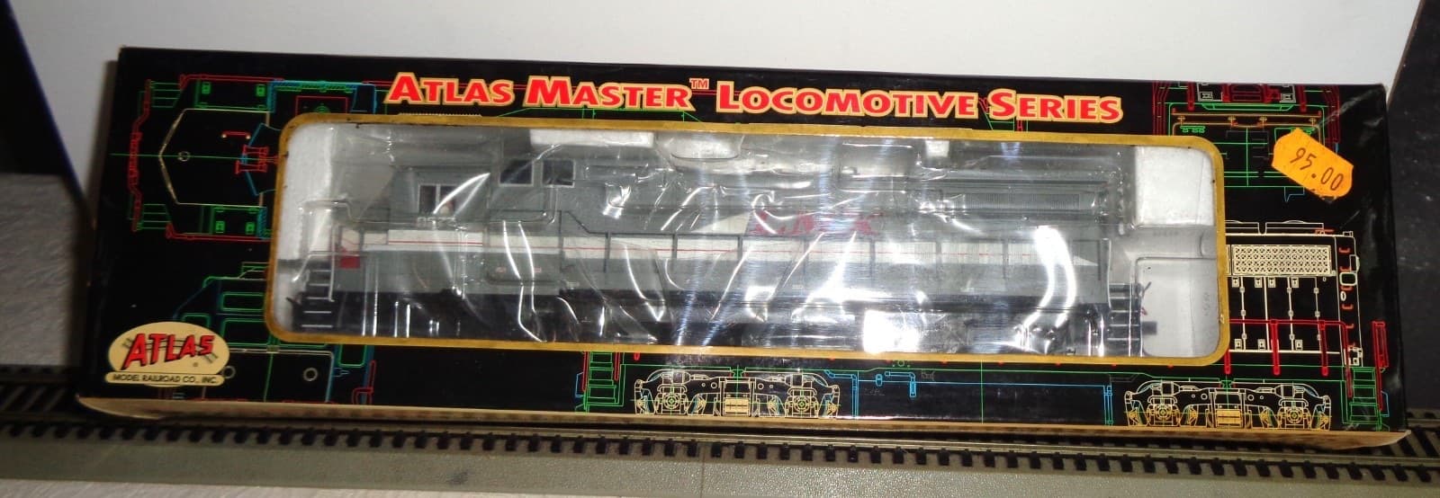 黒い箱の窓に入った灰色のディーゼル機関車の模型。箱の上に「ATLAS MASTER LOCOMOTIVE SERIES」の文字と、配線の図のような模様