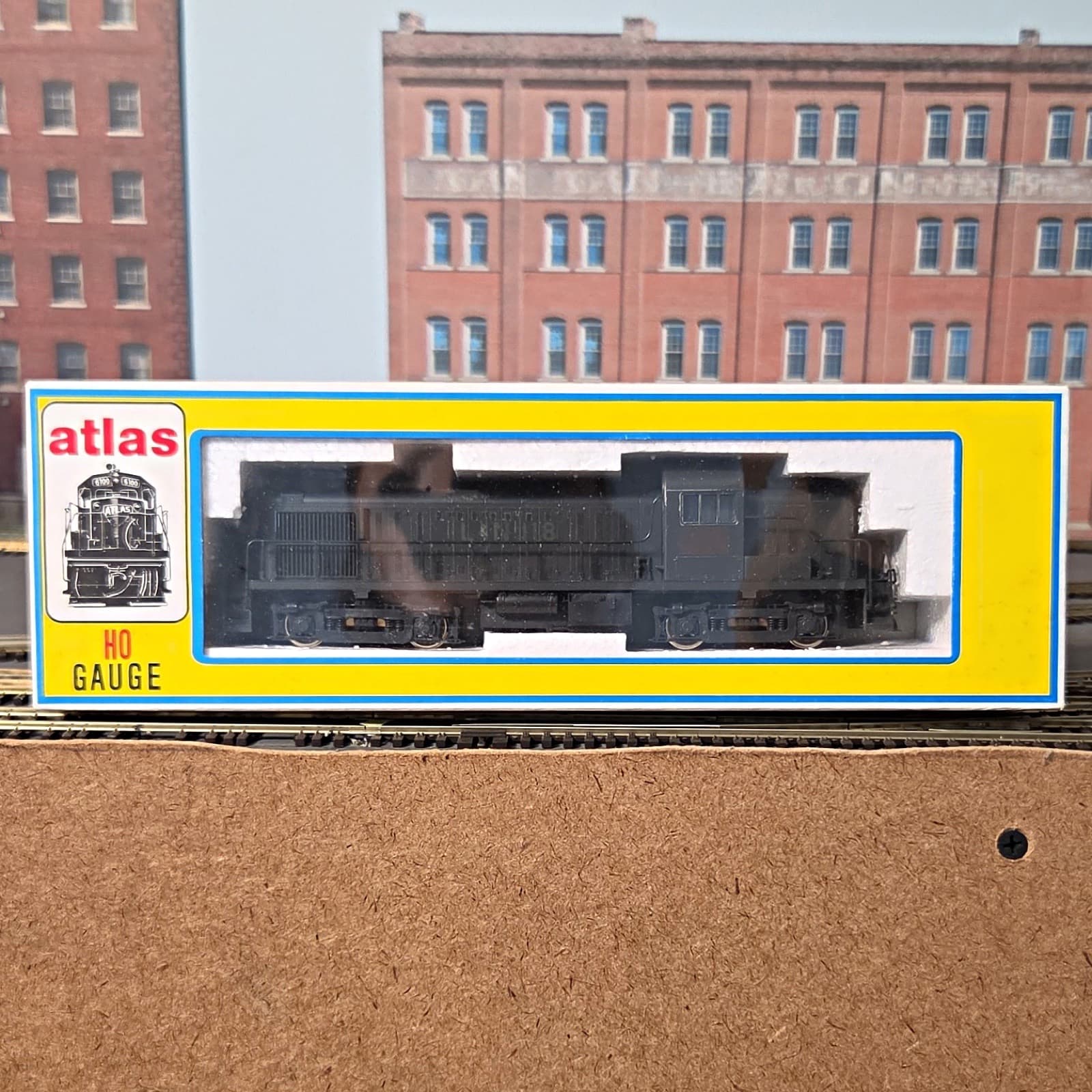 黄色い箱の窓に入った黒いディーゼル機関車の模型。箱の左に「atlas」「HO GAUGE」の文字と機関車の絵