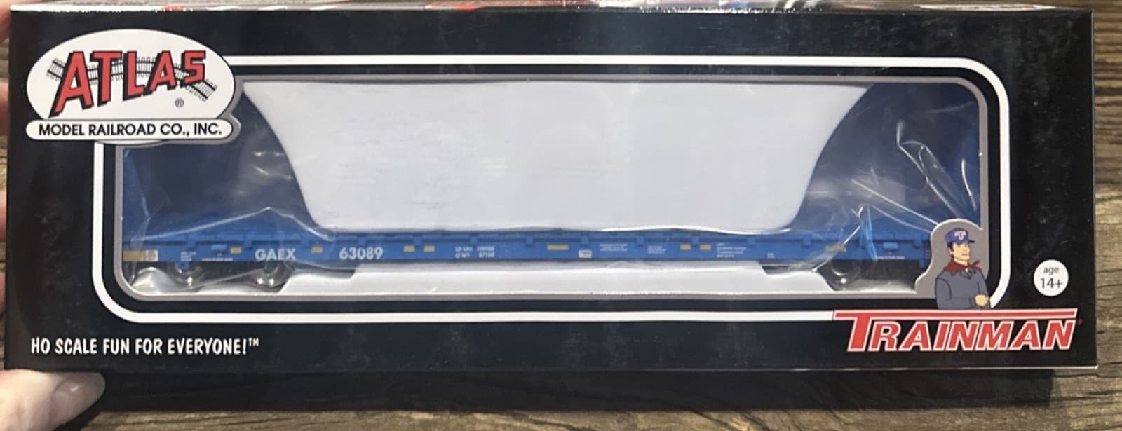 黒い枠の箱の窓に入った青い長物車の模型。箱の右下に赤い「TRAINMAN」の文字