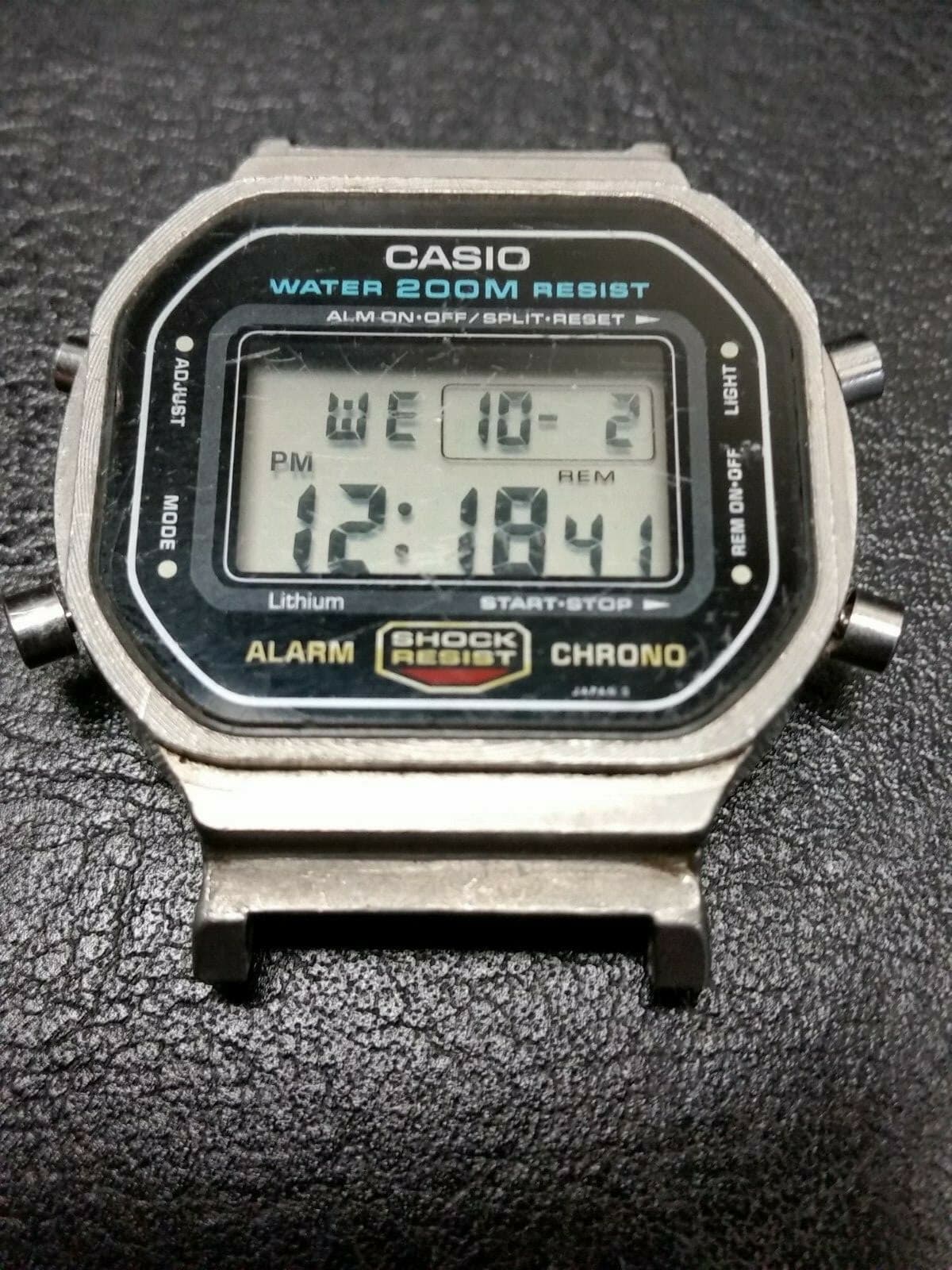 バンドとベゼルを外した、銀色の金属の角型のケースの時計。液晶の上に「WATER 200M RESIST」、左下に「Lithium」の文字がある