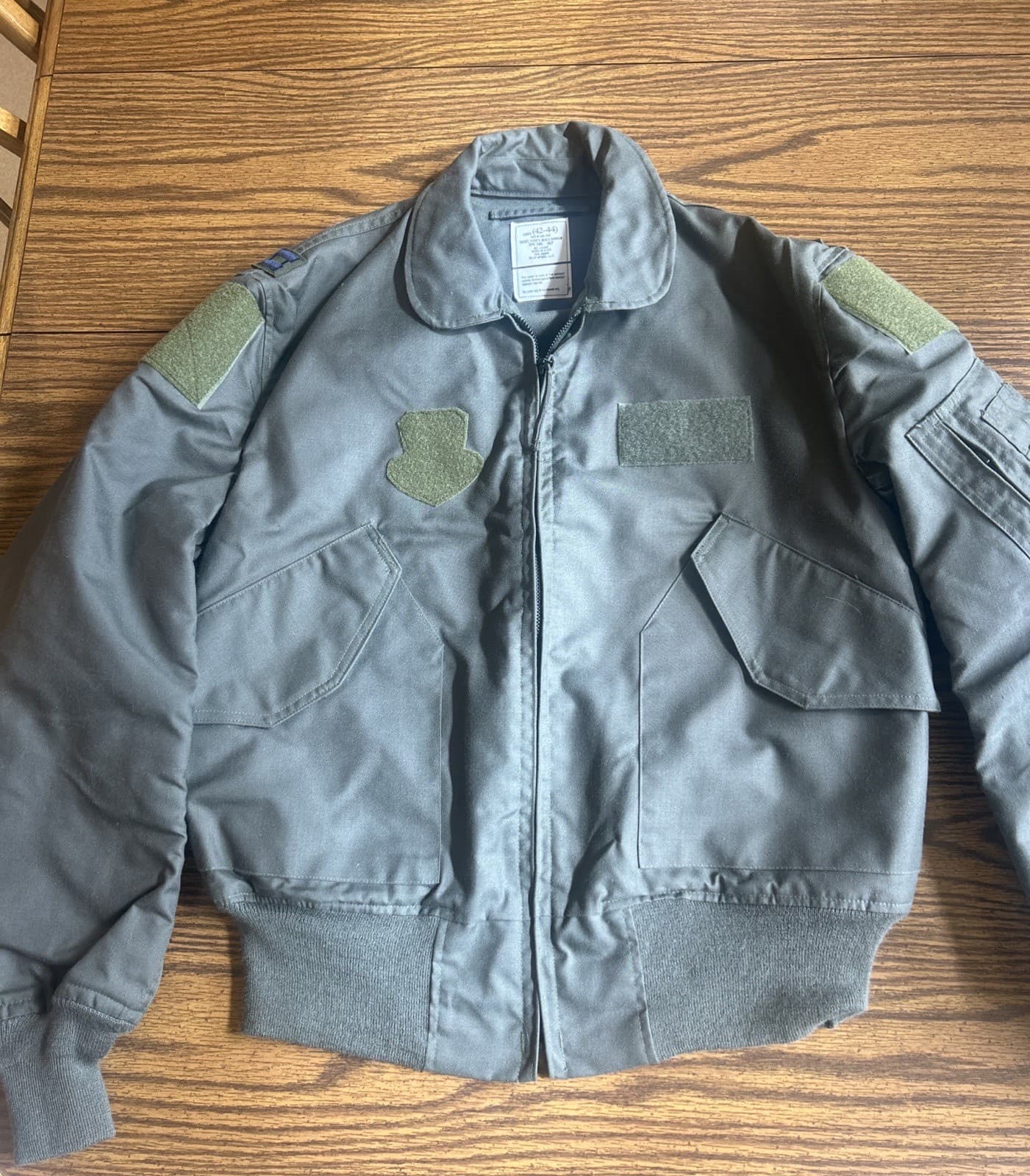 木の机に広げた、つやの少ない緑の飛行ジャケットの前面。肩と胸に面ファスナーが付いている