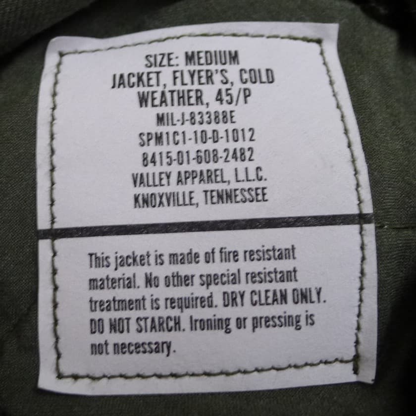 白い1枚のラベル。上に「SIZE: MEDIUM」「JACKET, FLYER'S, COLD WEATHER, 45/P」「MIL-J-83388E」「SPM1C1-10-D-1012」「VALLEY APPAREL, L.L.C.」、線の下に「DRY CLEAN ONLY」を含む手入れの文
