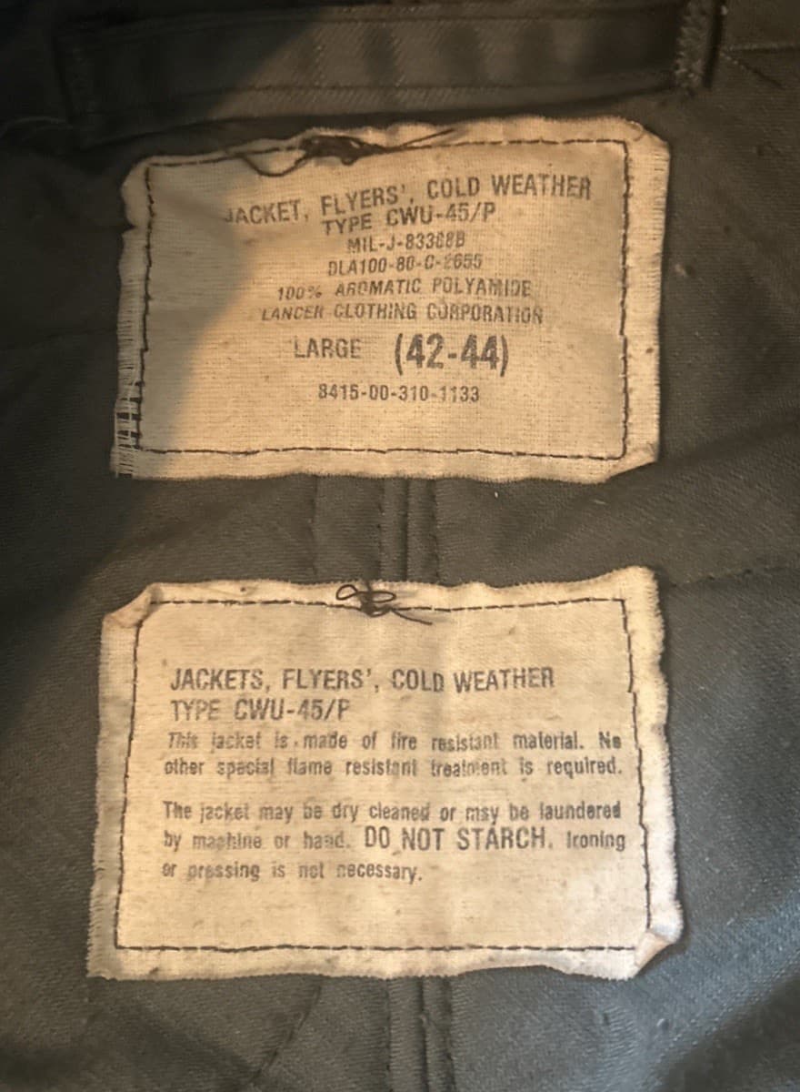 上下に2枚の布のラベル。上は「TYPE CWU-45/P」「MIL-J-83388B」「DLA100-80-C-2655」「LANCER CLOTHING CORPORATION」「LARGE (42-44)」、下は洗い方の注意の文