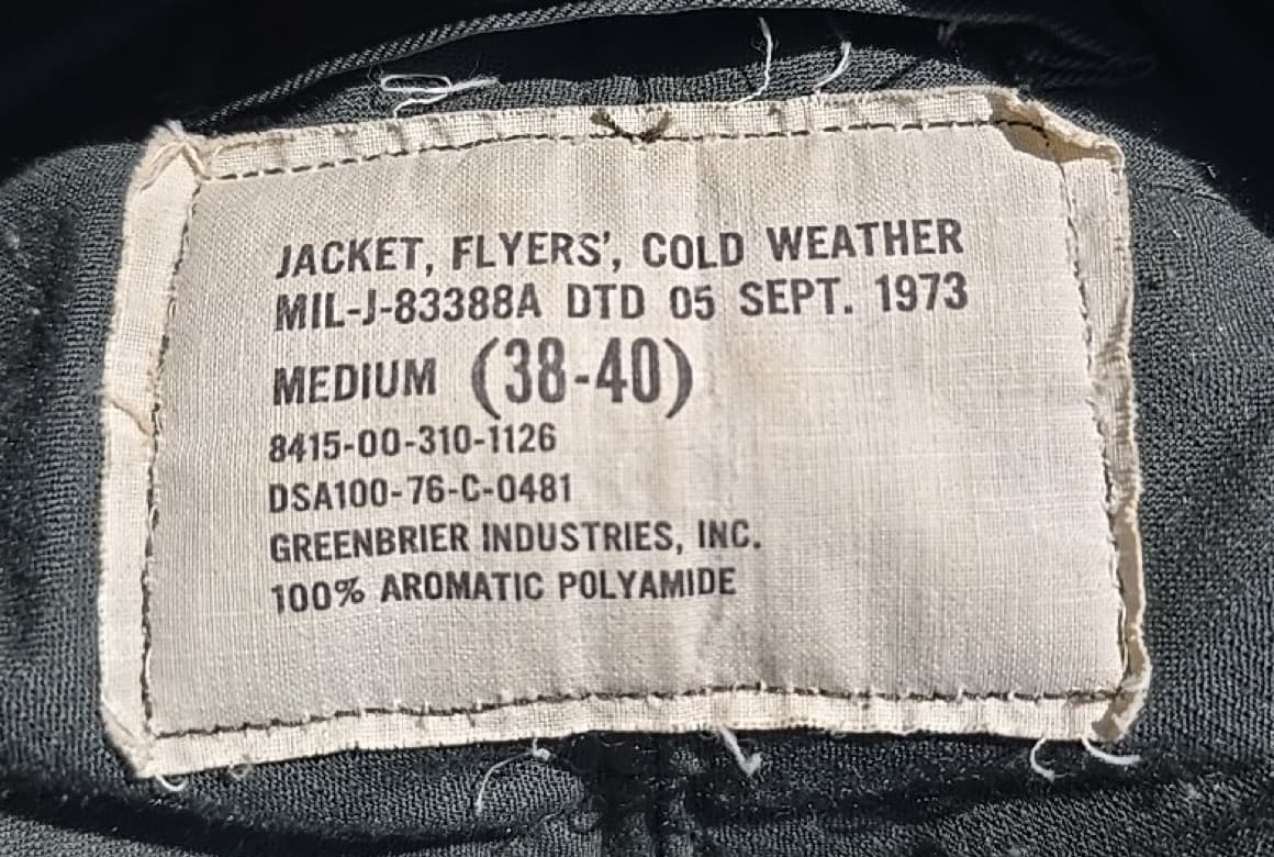 白い布のラベル。「JACKET, FLYERS', COLD WEATHER」「MIL-J-83388A DTD 05 SEPT. 1973」「MEDIUM (38-40)」「DSA100-76-C-0481」「GREENBRIER INDUSTRIES, INC.」などの文字が並ぶ