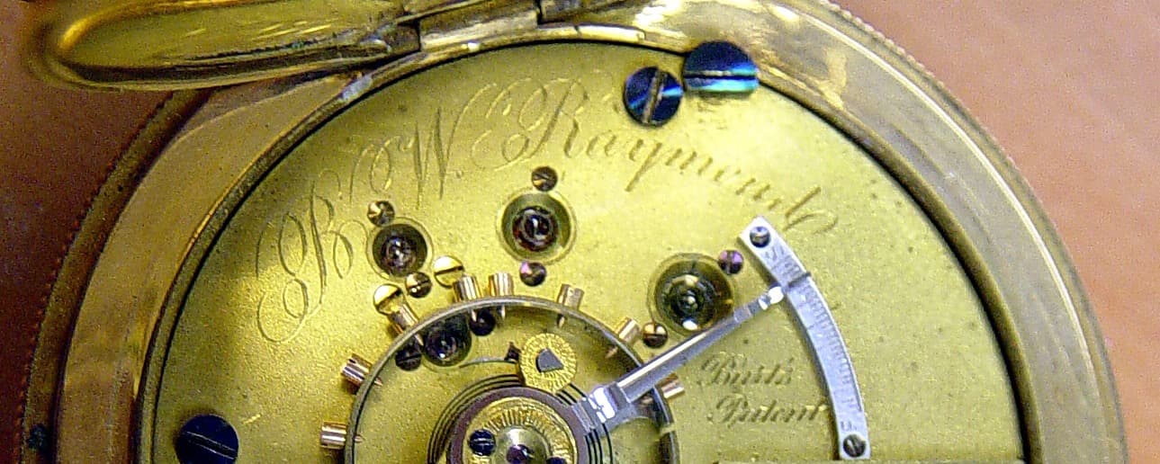懐中時計の機械の受け板の一部。筆記体の「B.W. Raymond」の彫りと「Burt's Patent」の文字、てんぷ、緩急針の目盛りが横長に写る