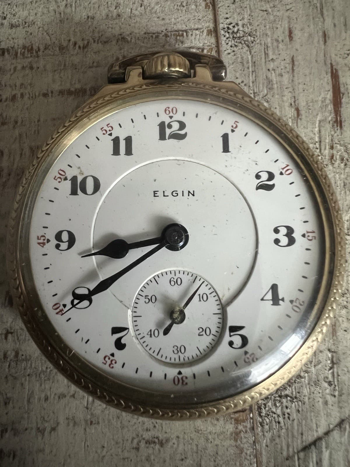 白い木の上に置いた金色のケースの懐中時計。白い文字盤に黒いアラビア数字と「ELGIN」の文字、下に秒針の小さな文字盤がある