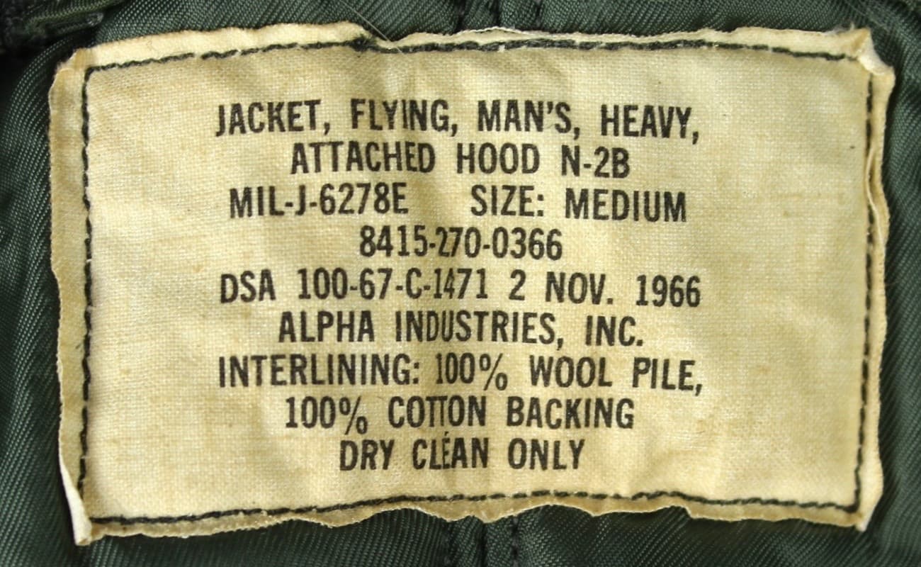 緑の裏地に縫い付けた生成りのタグ。品名、規格番号 MIL-J-6278E、在庫番号、DSA で始まる契約番号と1966年の日付、アルファ社の名前、中綿の素材が黒で印字されている