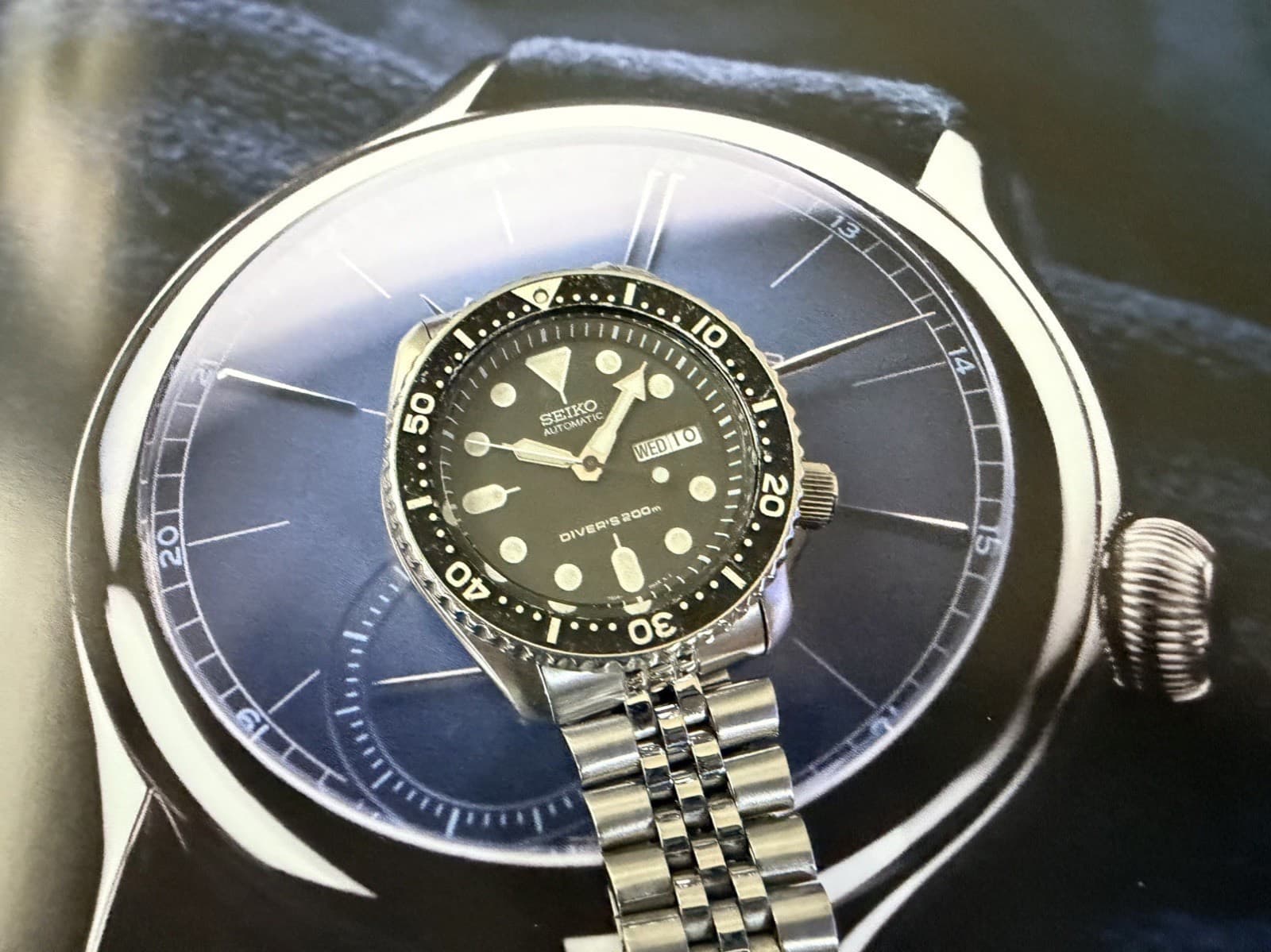 青い時計の文字盤を印刷した紙の上に置いた、5連の金属のブレスレットの黒い文字盤のダイバーズ