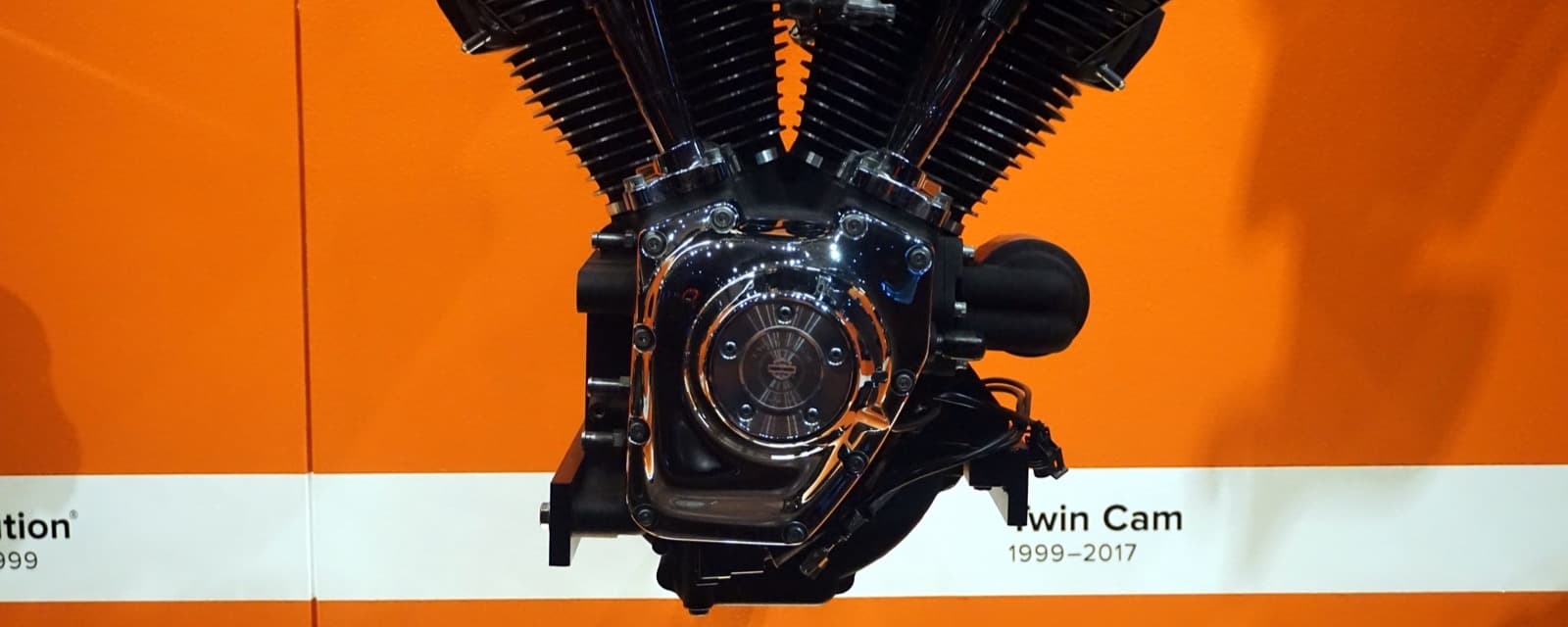 オレンジの壁の前に展示された黒い V 型2気筒のエンジンの下半分。右側に丸いカムのカバーがあり、下の白い帯に「Twin Cam 1999–2017」の札