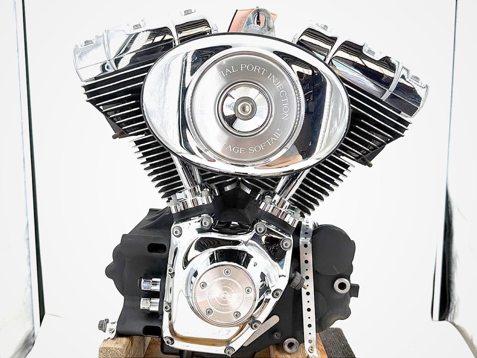 白い布の前の木の台に載せたV型2気筒のエンジン。楕円のエアクリーナーに「…TIAL PORT INJECTION」「…ITAGE SOFTAIL」の文字