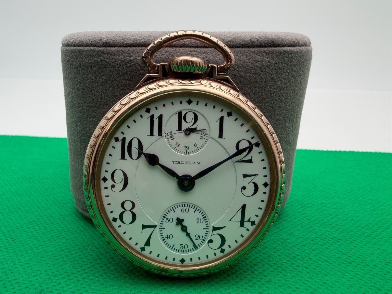 灰色の台に立てた金色の懐中時計。白い文字盤の12時の下と6時の位置に、小さな目盛りが2つある