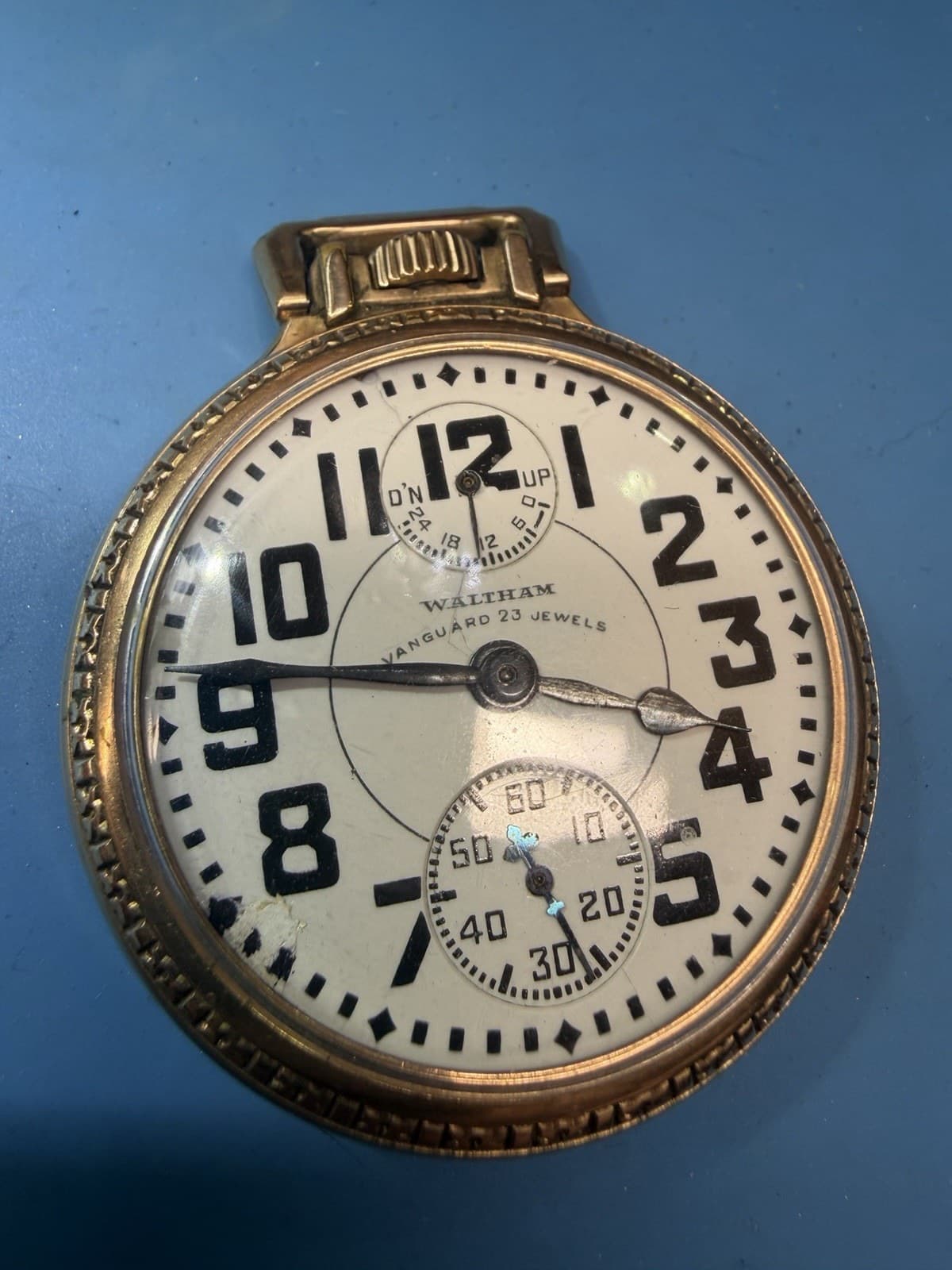 青い背景の前の金色の懐中時計。白い文字盤に黒い太いアラビア数字があり、12時の下に小さな目盛りの窓がある