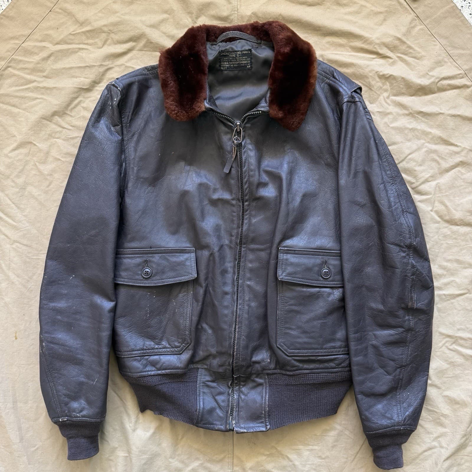 生成りの布の上に置いた、青みがかって写った濃い色の革の上着。茶の毛皮の襟と、胸の2つのフラップ付きのポケットがある